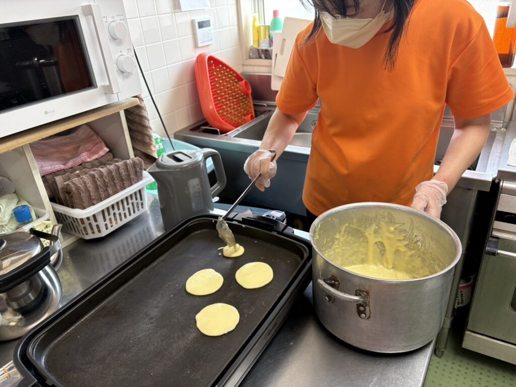 どら焼き作り。大きなボウルに入った生地を、お玉でホットプレートに丸く流し入れている調理風景。横には電子レンジや調理器具が並んでいる。